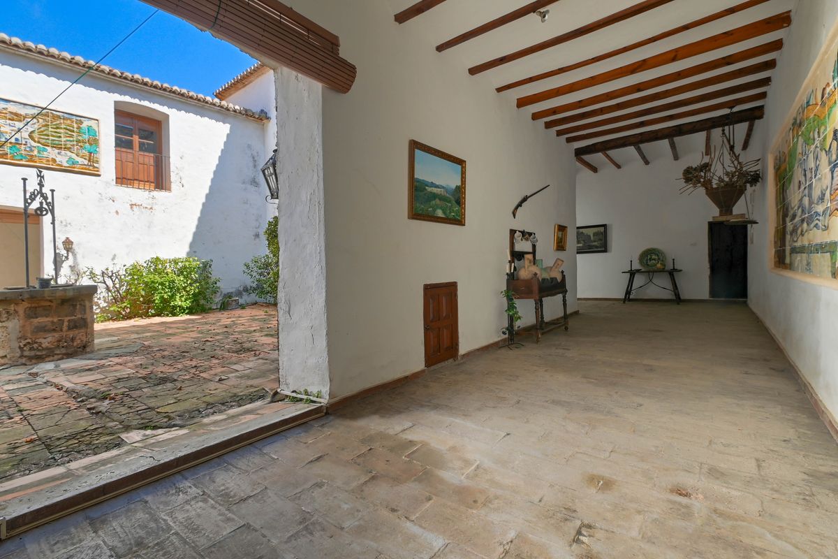 Covered porch with exposed wooden beams and stone flooring, featuring a traditional ceramic tile mural and views of a courtyard with an original stone well.