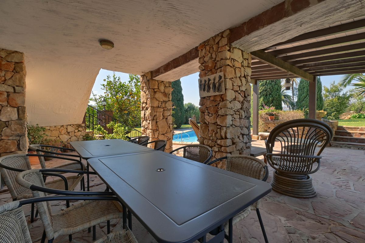 Covered terrace with dry stone pillars, outdoor dining area, and flagstone flooring overlooking the swimming pool and garden.