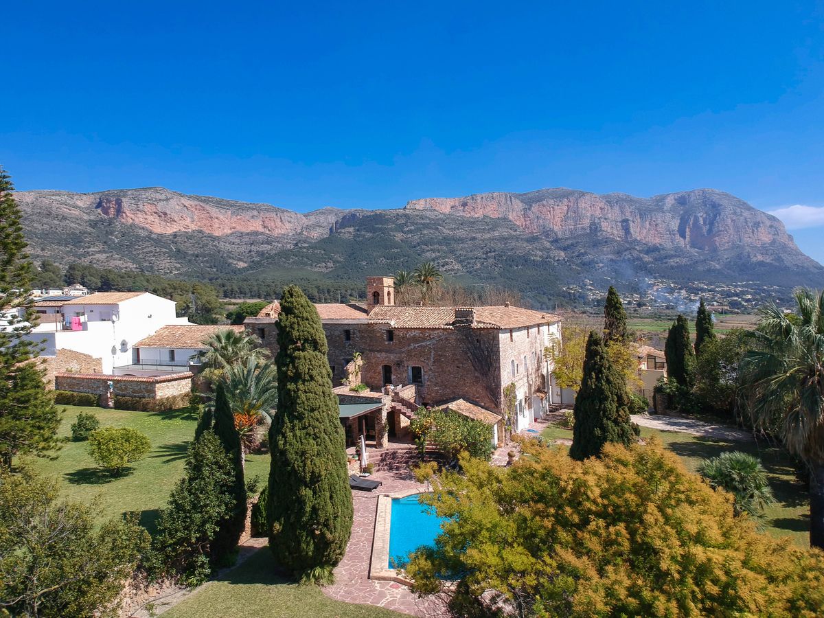 Elevated view of the historic stone finca featuring a swimming pool, manicured gardens with cypress trees, and the Montgó mountain range in the background.