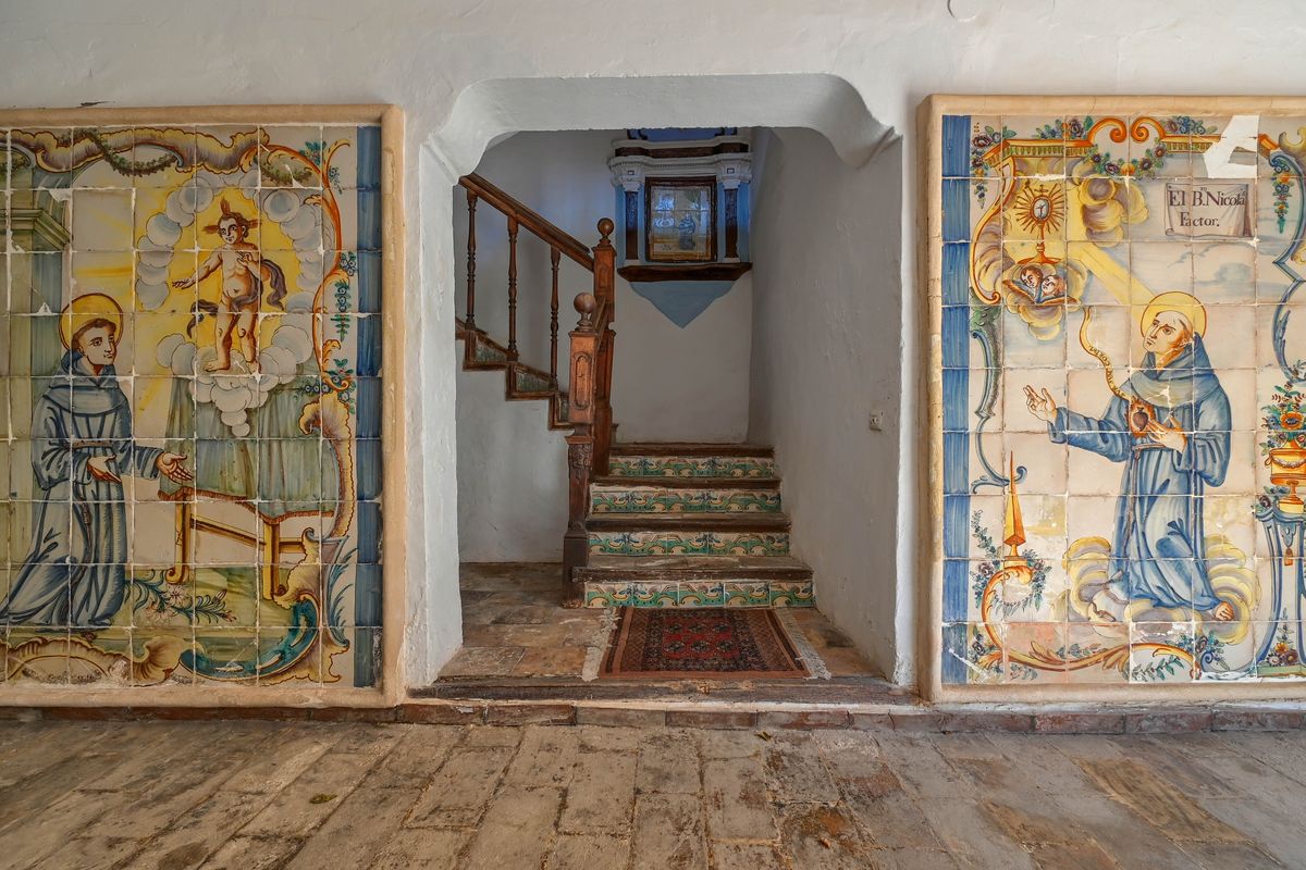 Entrance hall featuring historic hand-painted ceramic murals, thick white-washed walls, and a staircase with decorative tiled risers and a wooden banister.