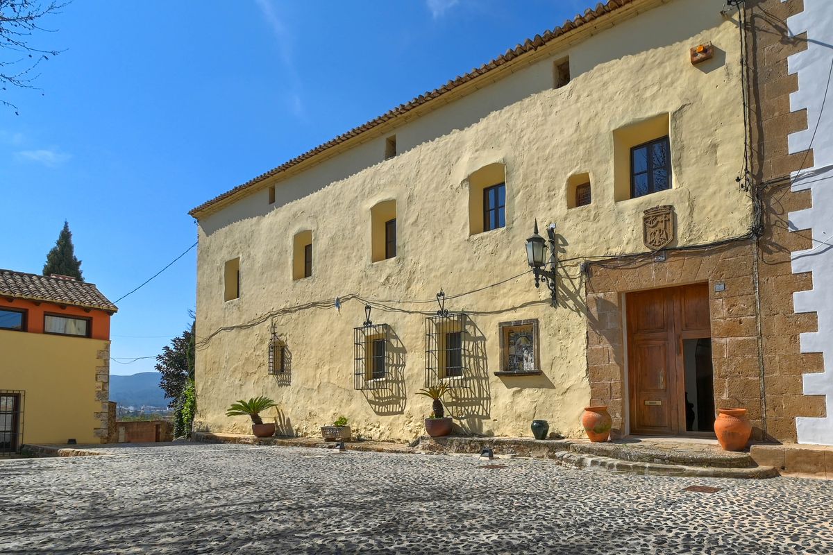 Historic facade with textured yellow walls, a Tosca stone portal, and a carved crest. The entrance is preceded by a traditional cobblestone courtyard and terracotta pots.