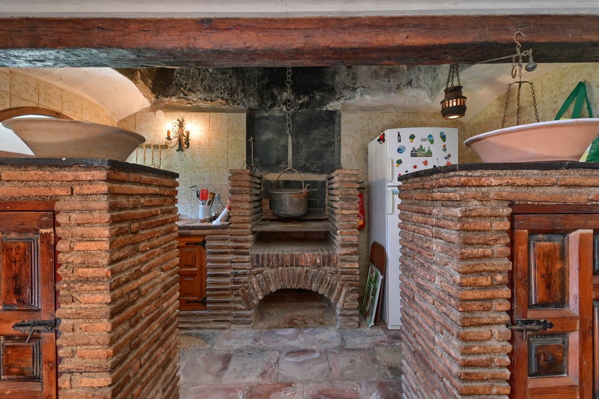 Historic kitchen area featuring a traditional open hearth with a hanging iron pot, exposed wooden beams, and rustic brickwork pillars.