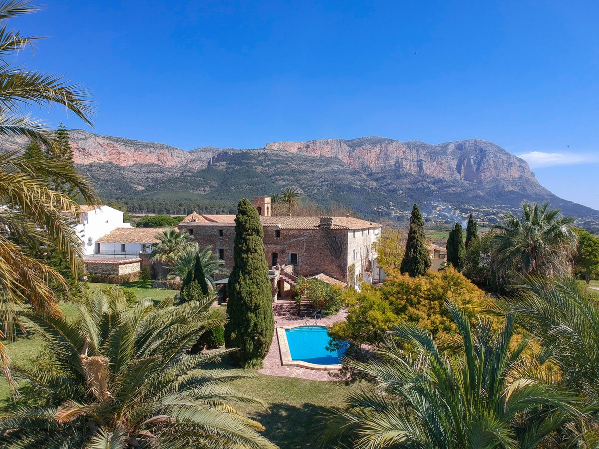 Historic stone finca featuring a swimming pool, lush Mediterranean gardens with palm trees, and panoramic views of the Montgó mountain range.