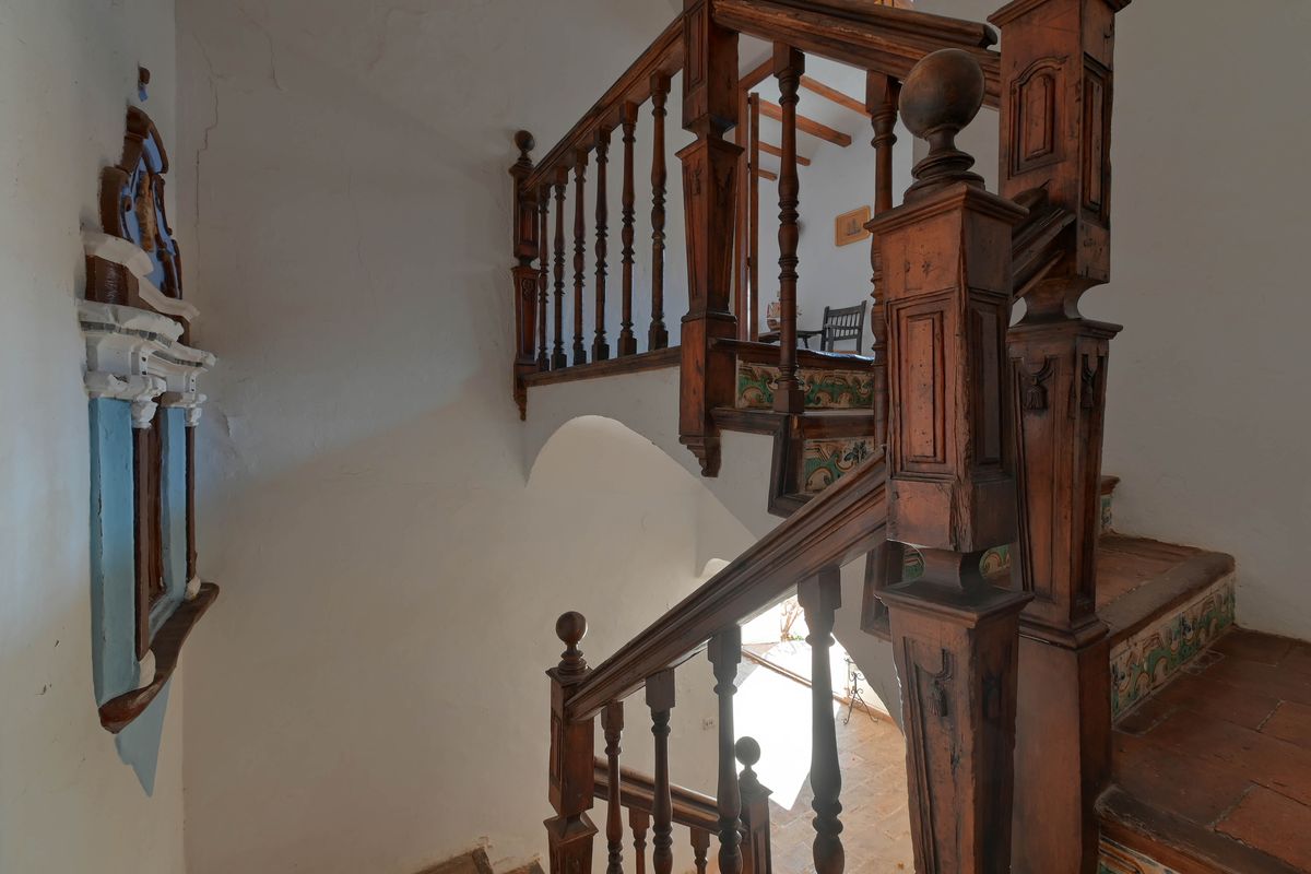 Historic wooden staircase featuring carved balustrades, terracotta steps with decorative ceramic risers, and white plastered walls.