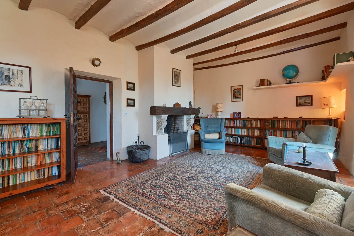 Living room featuring exposed dark wood ceiling beams, a classic fireplace with a timber mantel, terracotta tile flooring, and integrated bookshelves.