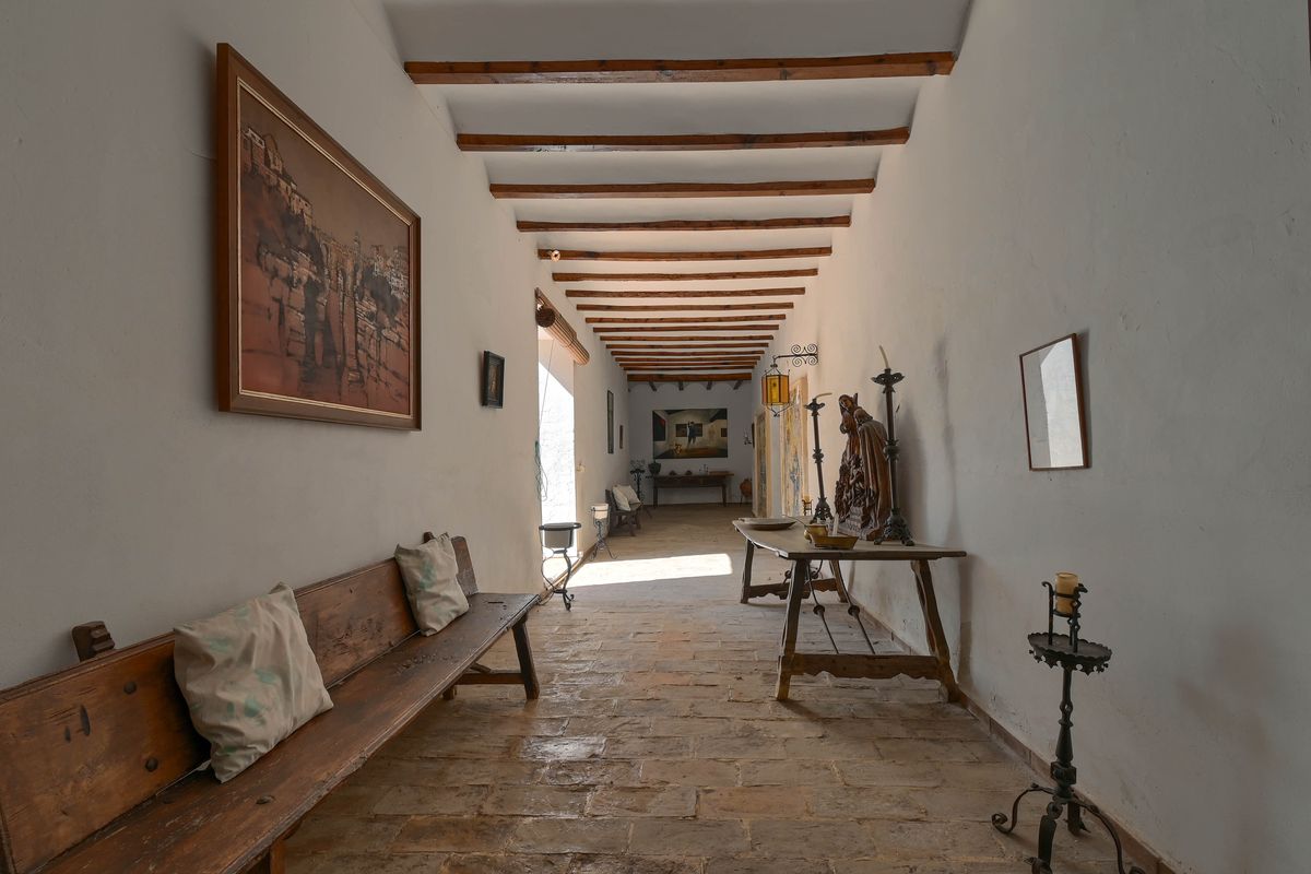 Long hallway featuring exposed timber beams, traditional stone tile flooring, and curated antique wooden furniture against white walls.