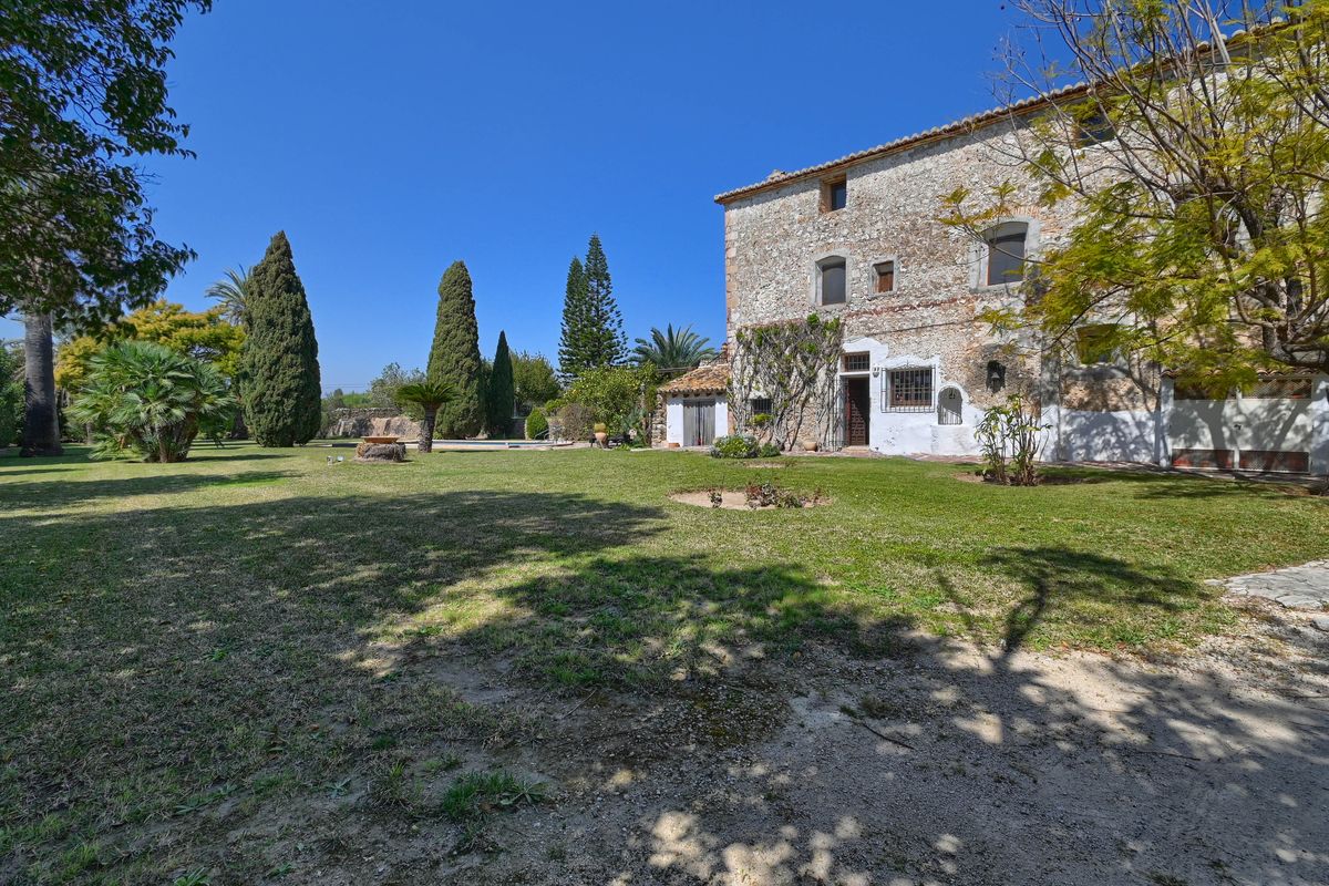 Natural stone facade of the historic finca surrounded by expansive lawn gardens with cypress and palm trees under a clear sky.