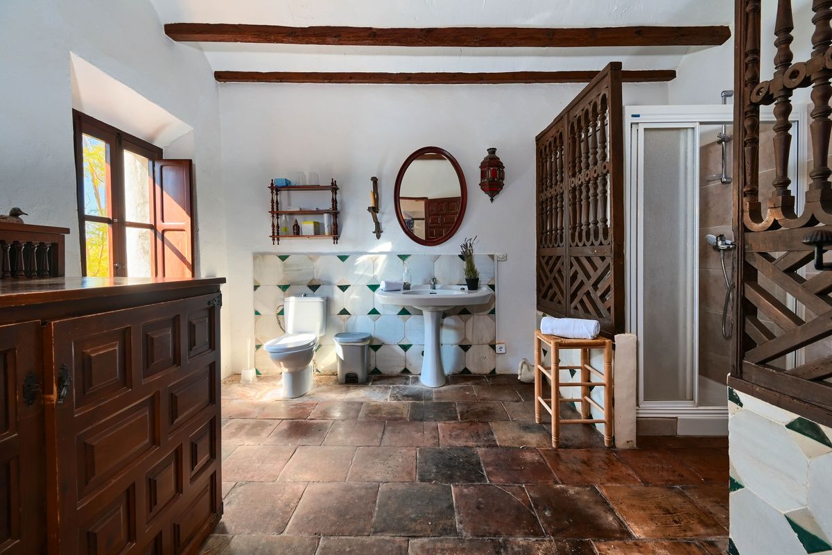 Rustic bathroom featuring terracotta floors, exposed ceiling beams, and a carved wooden screen. Includes a pedestal sink against a decorative tiled wall.