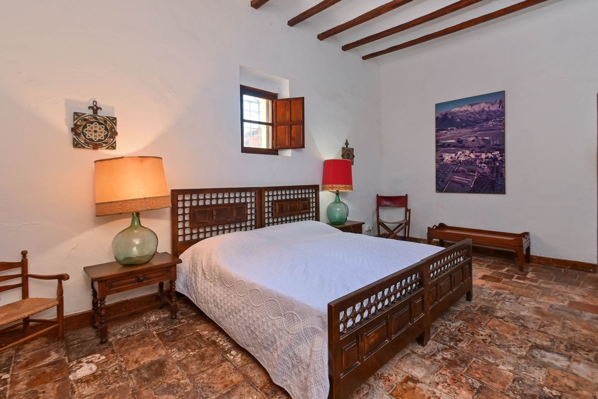 Rustic bedroom featuring authentic terracotta floors, exposed wooden ceiling beams, and a carved dark wood bed frame. Traditional shutters and demijohn lamps enhance the historic character.