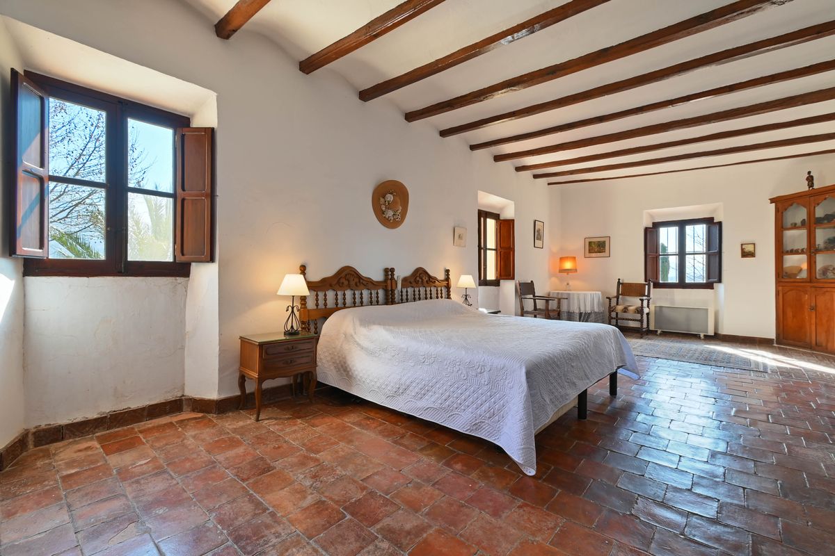Rustic bedroom featuring exposed dark wood ceiling beams, authentic terracotta floor tiles, and traditional carved wooden furniture.