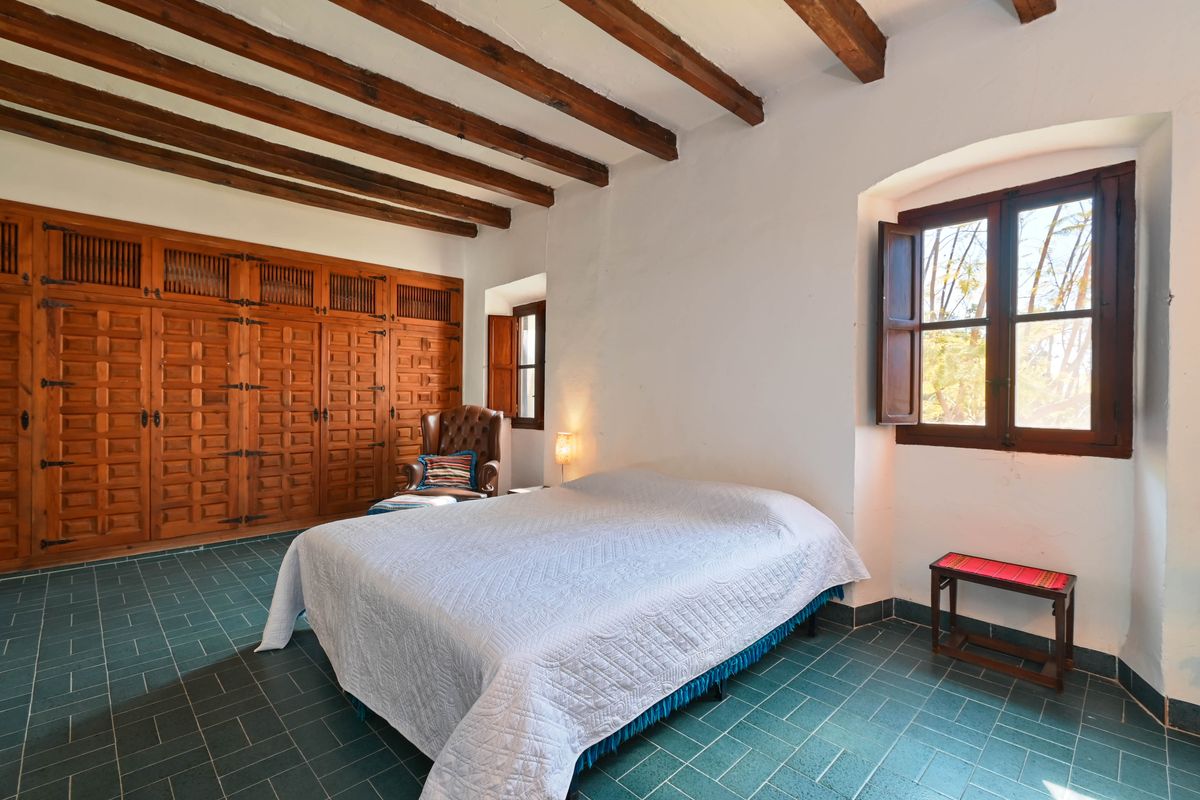 Rustic bedroom featuring exposed wooden ceiling beams, built-in carved wood wardrobes, green tiled flooring, and deep-set windows with traditional shutters.