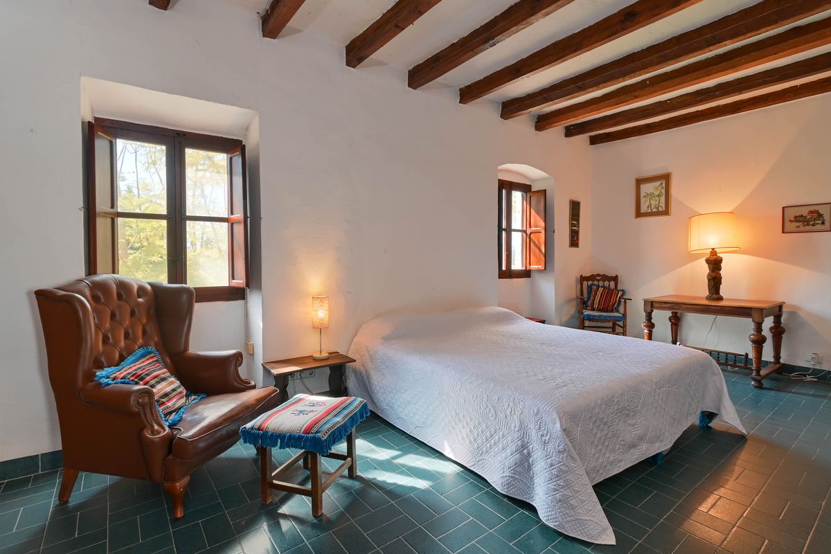 Rustic bedroom featuring exposed wooden ceiling beams, green tiled flooring, and a classic leather wingback chair by traditional shuttered windows.