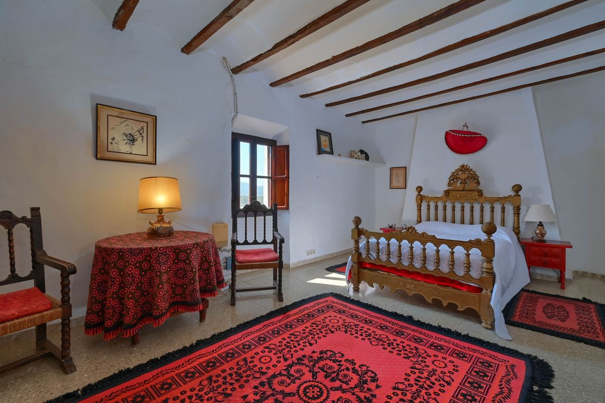 Rustic bedroom featuring original wooden ceiling beams, a hand-carved bed frame, and decorative red rugs over stone flooring.