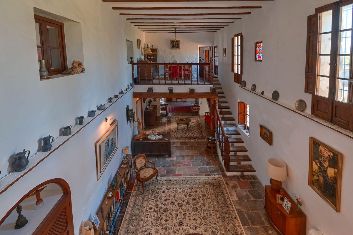 Spacious double-height hall featuring original terracotta floors, exposed timber beams, and a wooden staircase leading to a mezzanine gallery with rustic railings.