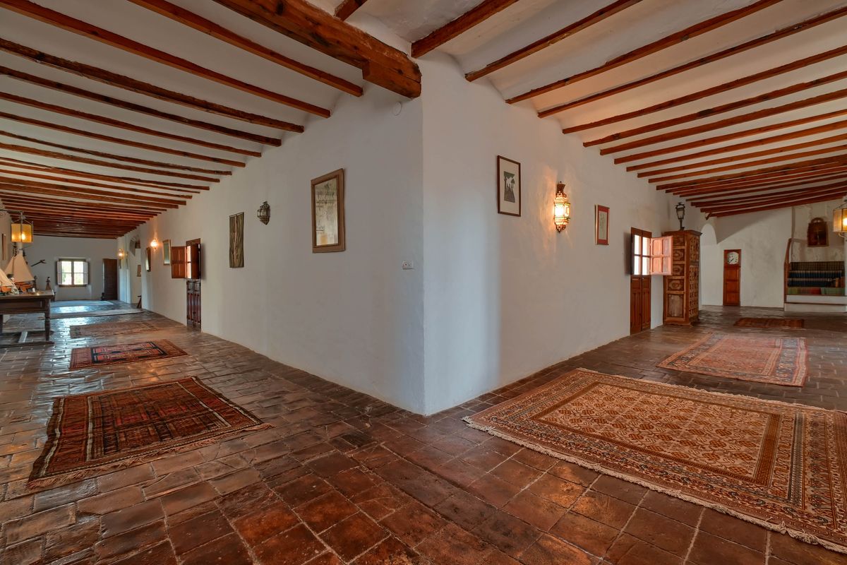 Spacious hallway featuring exposed wooden ceiling beams, original terracotta flooring, and traditional rugs under warm wall-mounted lanterns.