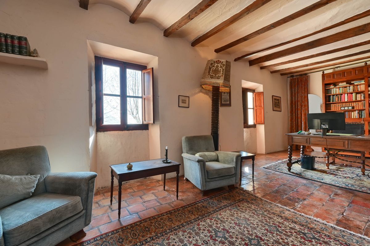 Study area featuring exposed wooden ceiling beams, terracotta floors, and deep-set windows. Includes an antique wooden desk and built-in library shelving.