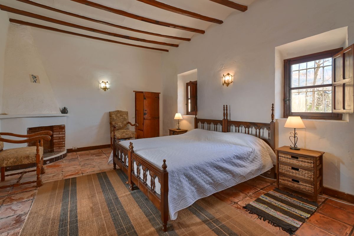 Traditional bedroom featuring exposed wooden ceiling beams, terracotta tile flooring, a built-in brick fireplace, and windows with original wooden shutters.