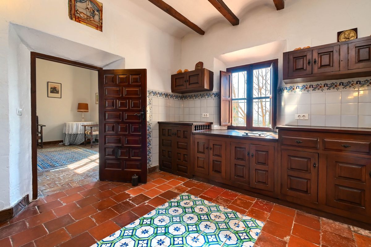 Traditional kitchen featuring dark wood cabinetry, terracotta flooring with decorative tile inlays, and exposed ceiling beams. A heavy wooden door leads to an adjacent room.