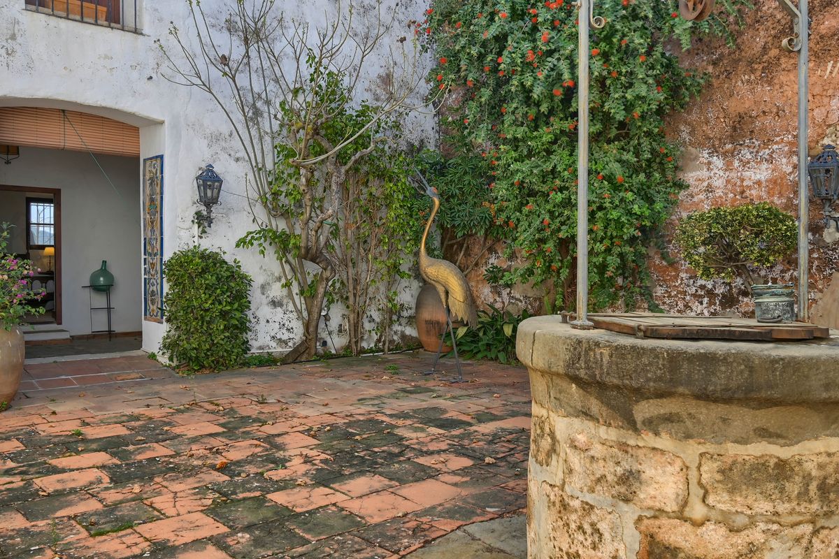 Traditional Mediterranean courtyard featuring an original stone well, weathered terracotta tiling, and a golden bird sculpture amidst lush climbing plants.