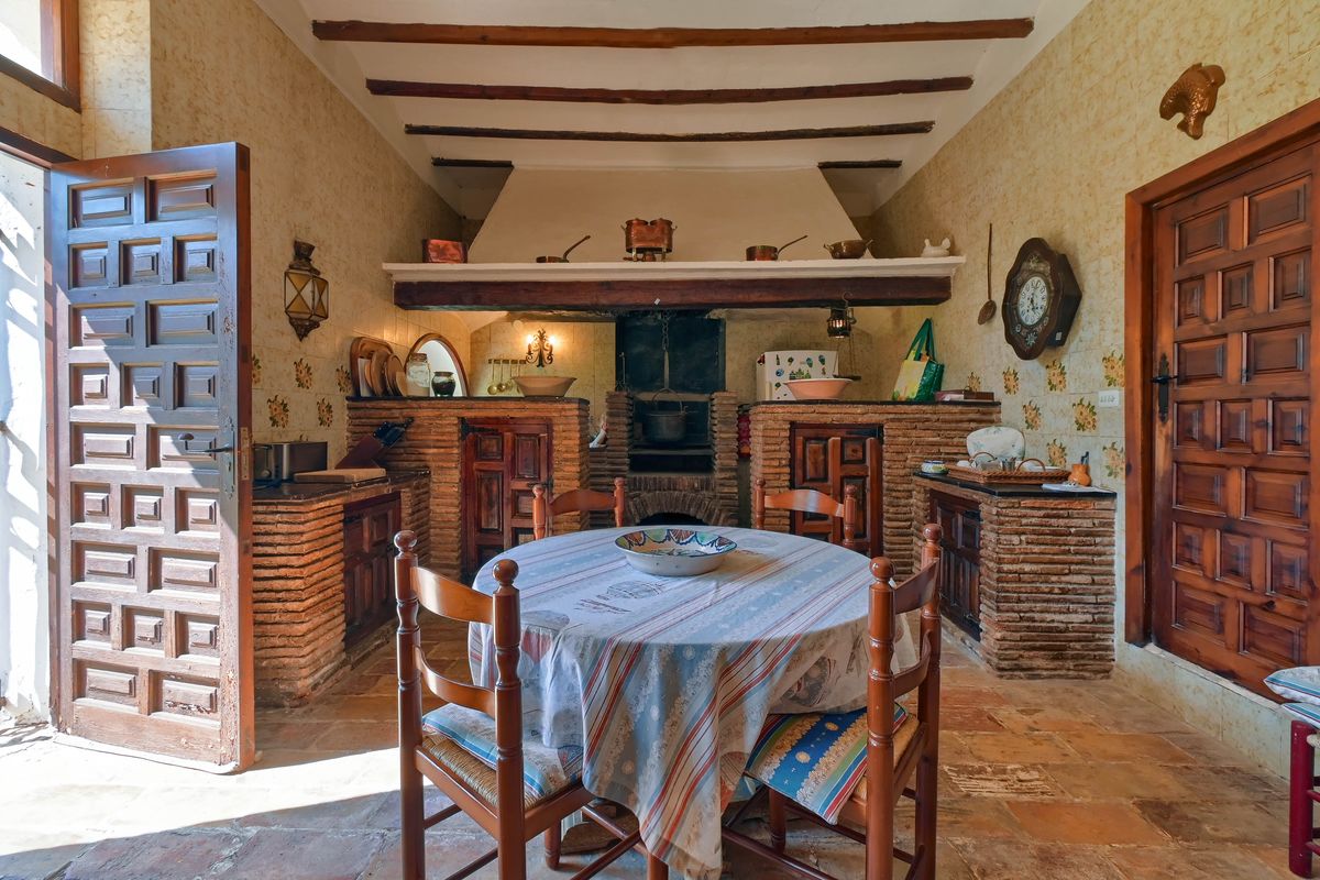 Traditional rustic kitchen featuring a large open brick hearth, exposed wooden ceiling beams, and original terracotta flooring with a central dining area.