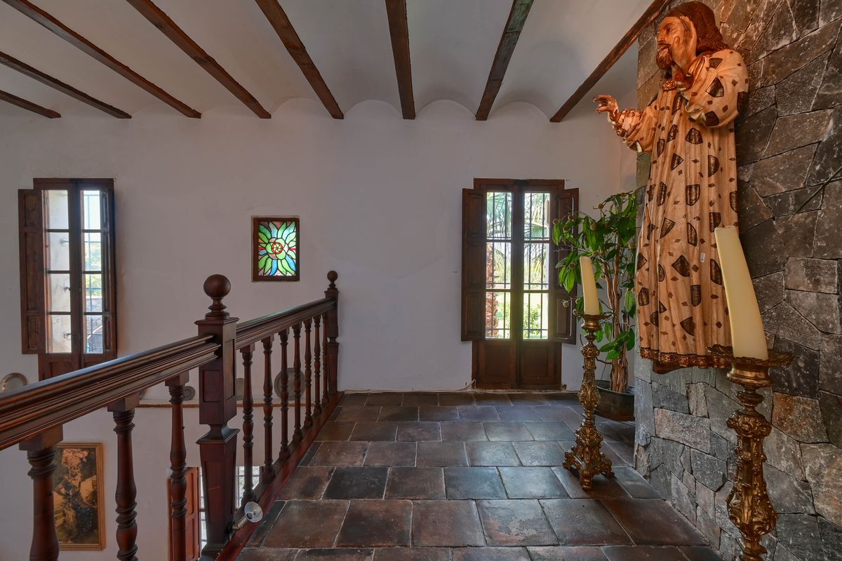Upper floor landing featuring terracotta tiles, exposed wooden ceiling beams, a carved wooden balustrade, and a stone accent wall with religious iconography.