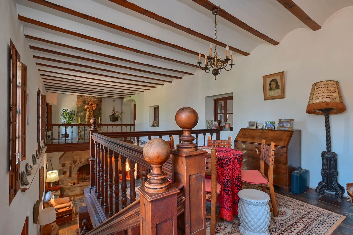 Upper mezzanine featuring exposed dark wood beams, a traditional wooden balustrade, and a study area with antique furniture overlooking the lower living room and stone fireplace.