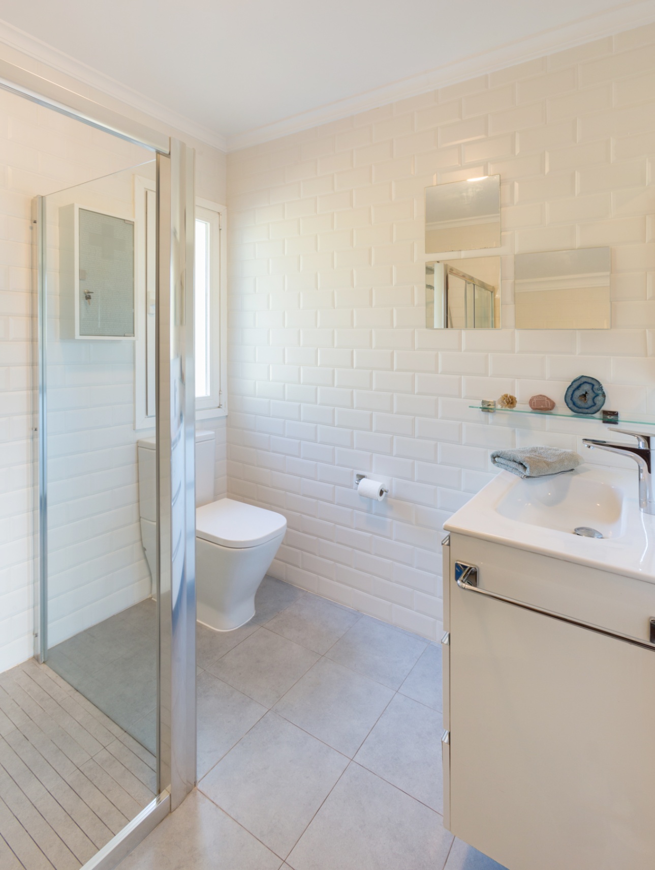 Bathroom featuring white subway wall tiles, a walk-in shower with glass partition, and a white vanity unit. Grey floor tiles and natural light enhance the space.