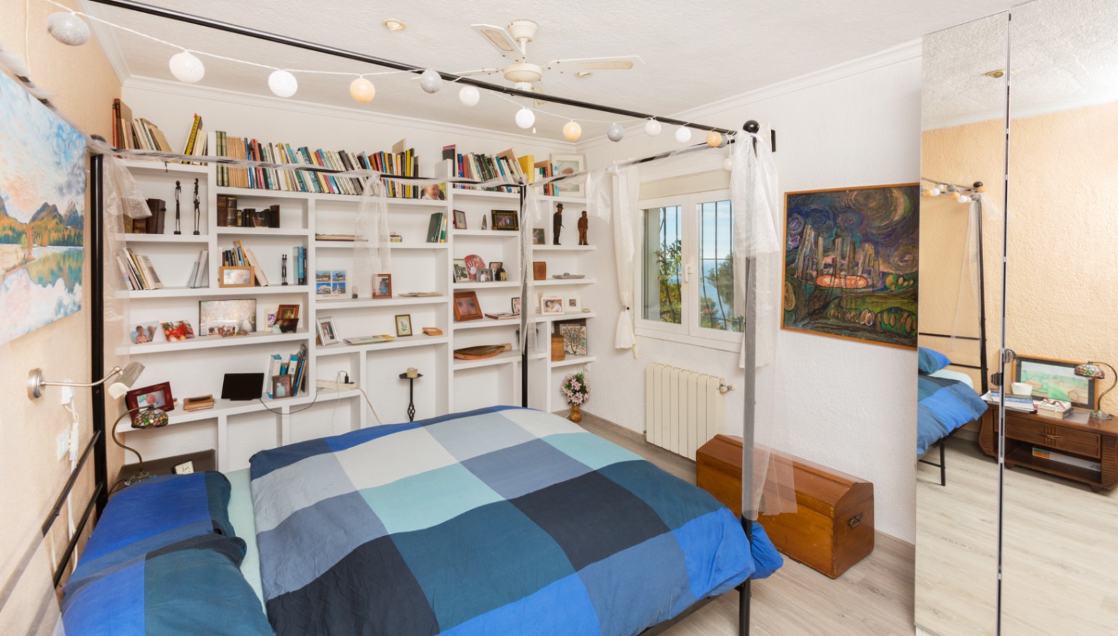 Bedroom with a black metal four-poster bed, integrated white wall shelving, and a large mirrored wardrobe. Features include a ceiling fan and a window with sea views.