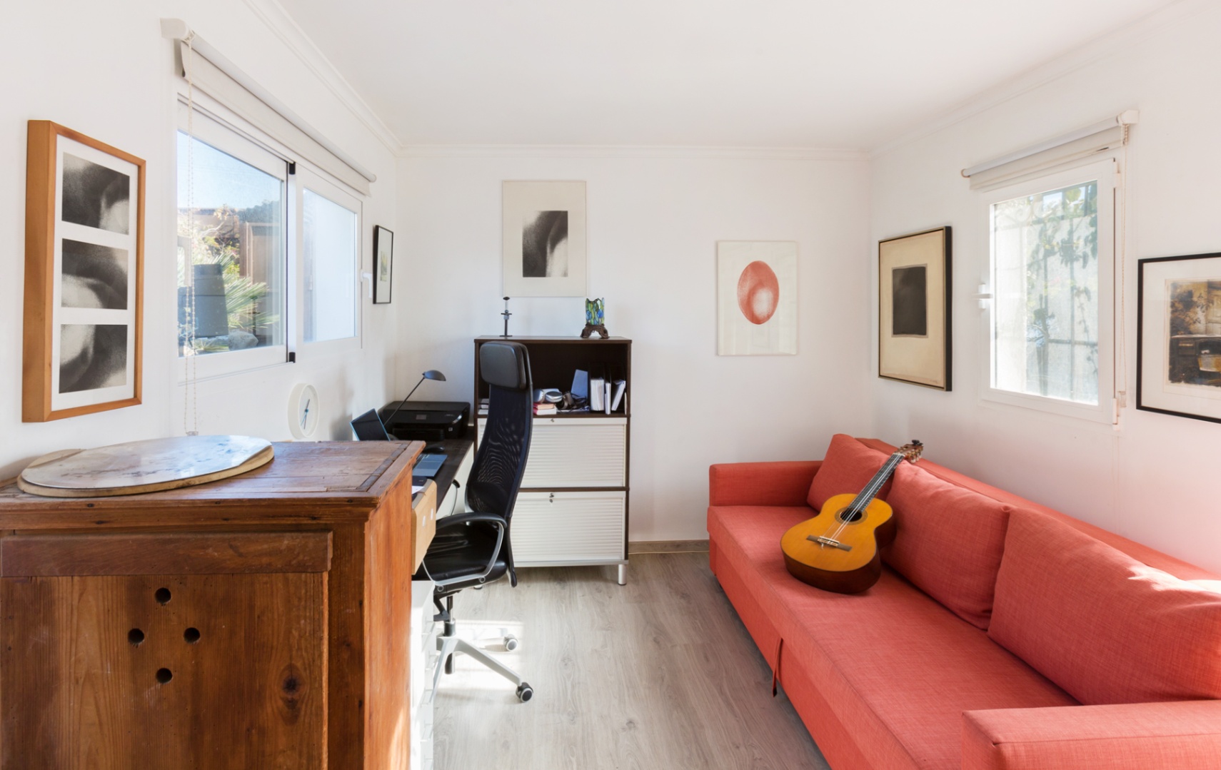 Bright home office with light wood flooring, featuring a red sofa, an ergonomic workspace, and natural light from dual-aspect windows.
