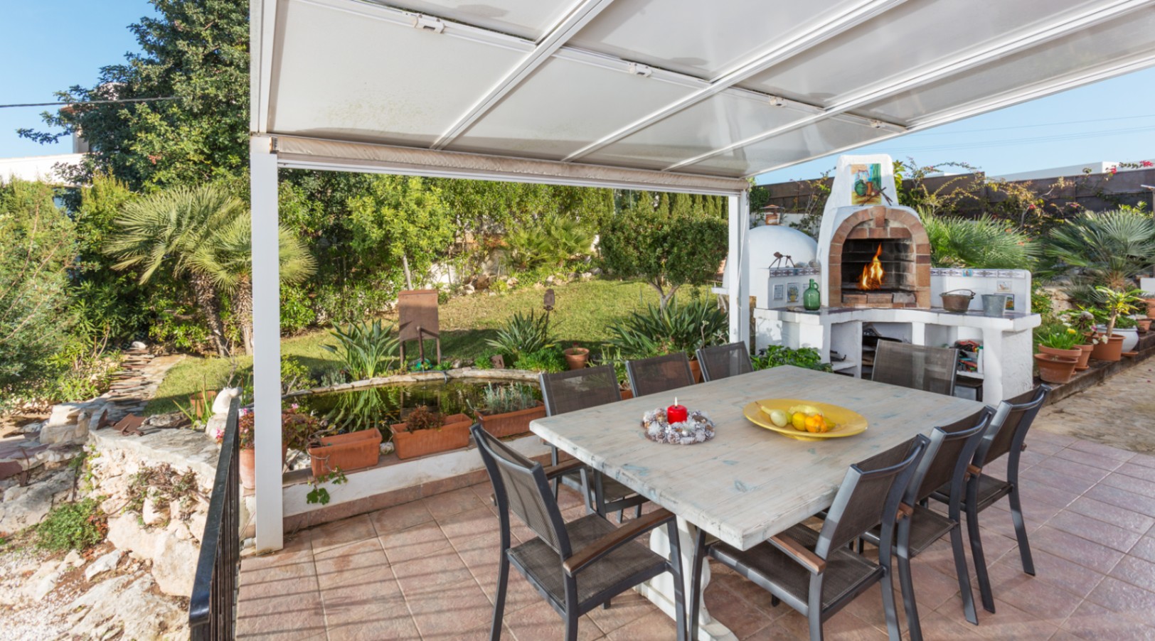 Covered outdoor terrace featuring a large wooden dining table, a traditional brick wood-fired oven, and views of the Mediterranean garden with a pond.