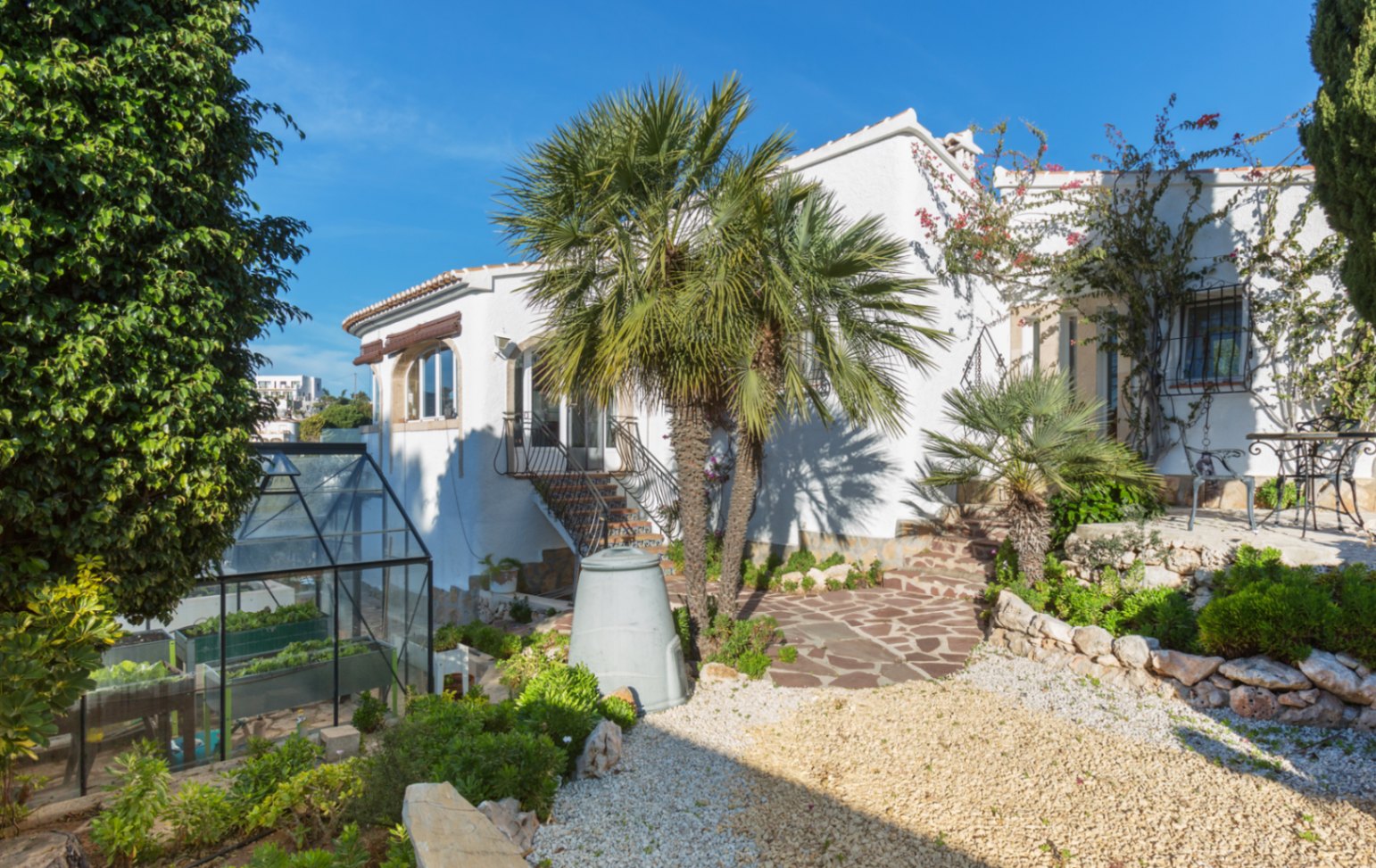 Exterior of a white Mediterranean villa featuring stone-paved paths, mature palm trees, and a glass greenhouse integrated into the landscaped garden.