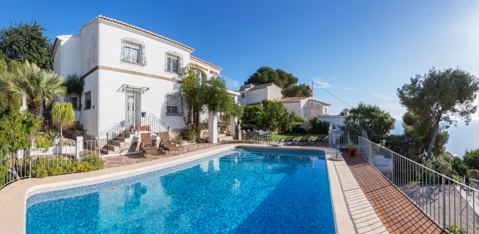 Exterior view of the Mediterranean villa featuring a large swimming pool, stone-paved terrace with sun loungers, and white facade with traditional ironwork overlooking the sea.