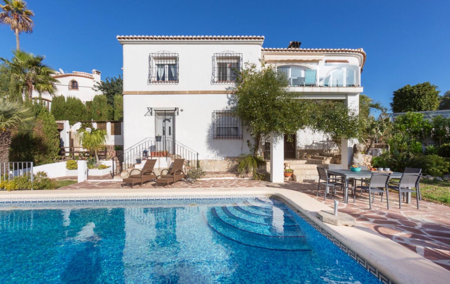 Exterior view of the villa featuring a private pool with Roman steps, a flagstone terrace with dining area, and a white facade with wrought iron bars.
