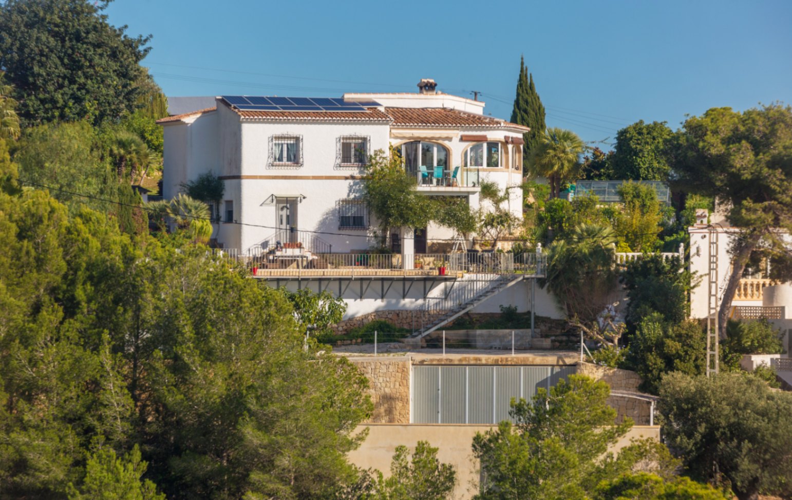 Multi-level Mediterranean villa featuring white facades, roof solar panels, and open terraces set against a backdrop of lush pine trees and stone walls.