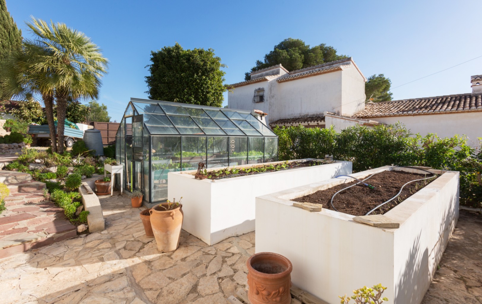 Outdoor garden area featuring a professional glass greenhouse and two large white raised planting beds with integrated irrigation systems on a stone terrace.
