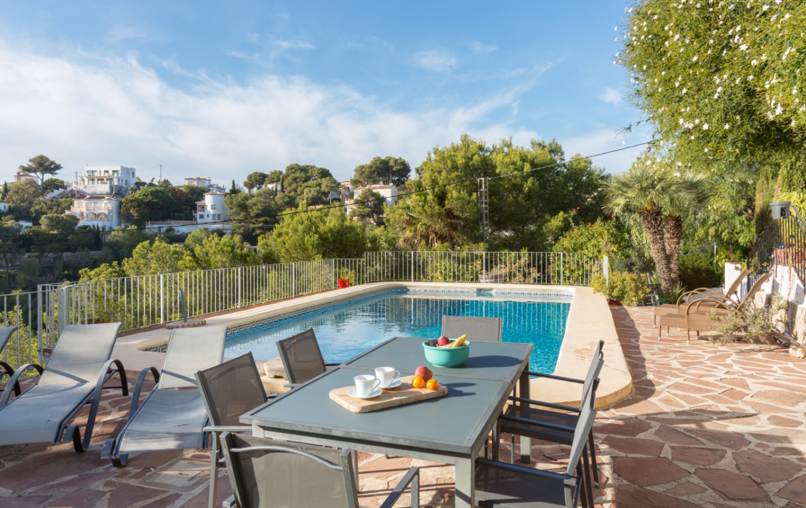 Outdoor terrace with irregular flagstone paving, a rectangular swimming pool, and a dining set, bordered by a metal railing and Mediterranean pine trees.