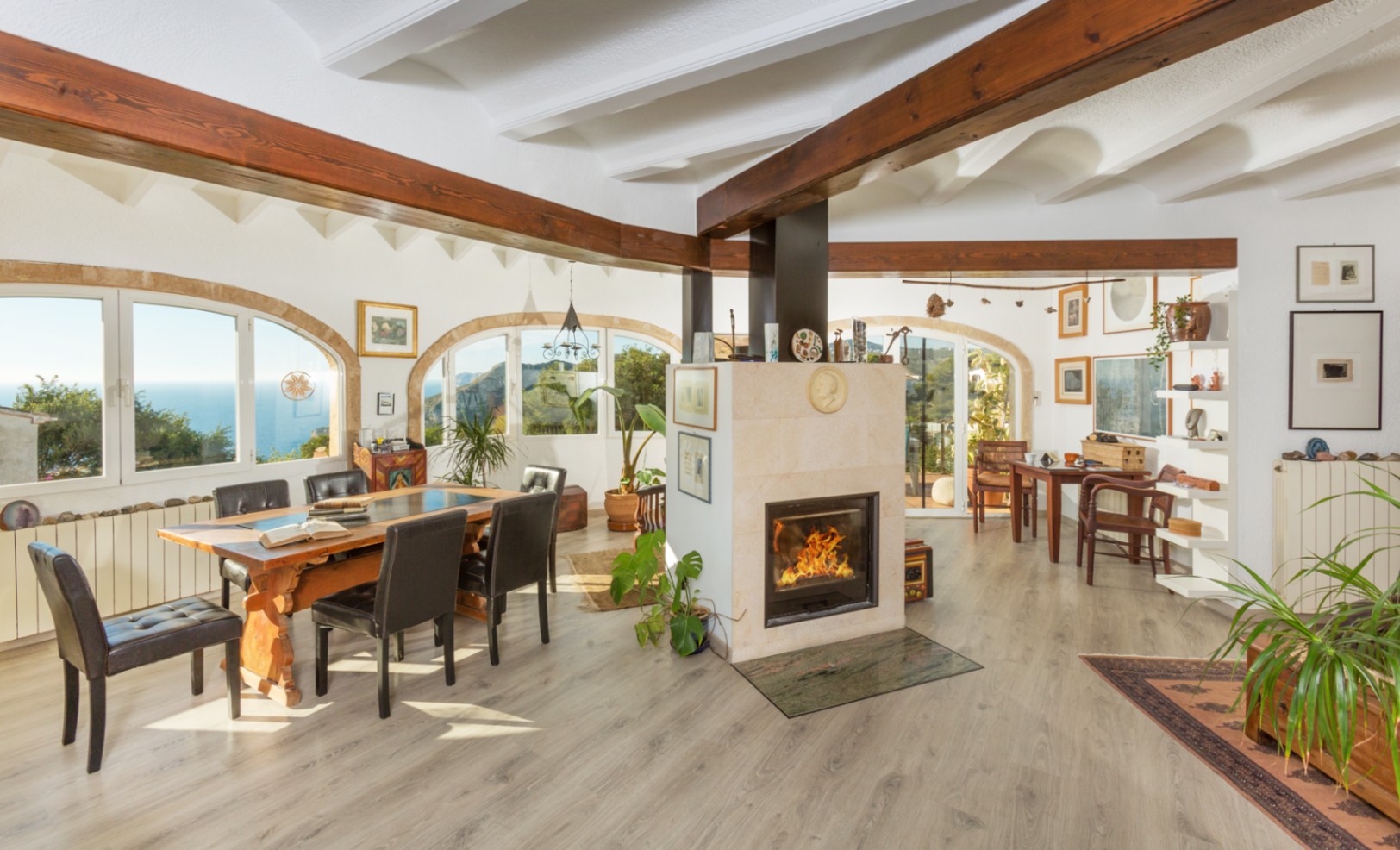 Spacious living and dining area featuring a central stone fireplace, exposed dark wood beams, and arched windows offering panoramic sea views.