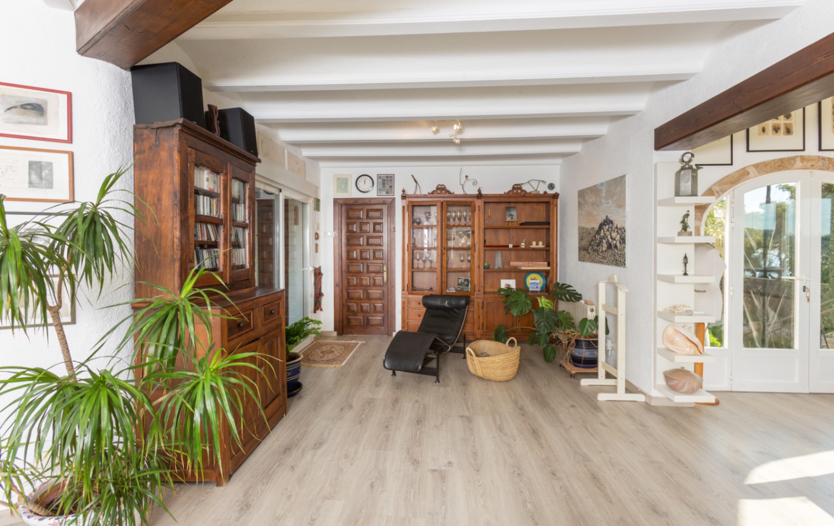 Spacious living area featuring white beamed ceilings, rustic wooden cabinetry, and light wood flooring. Natural light enters through glass doors with stone arch details.