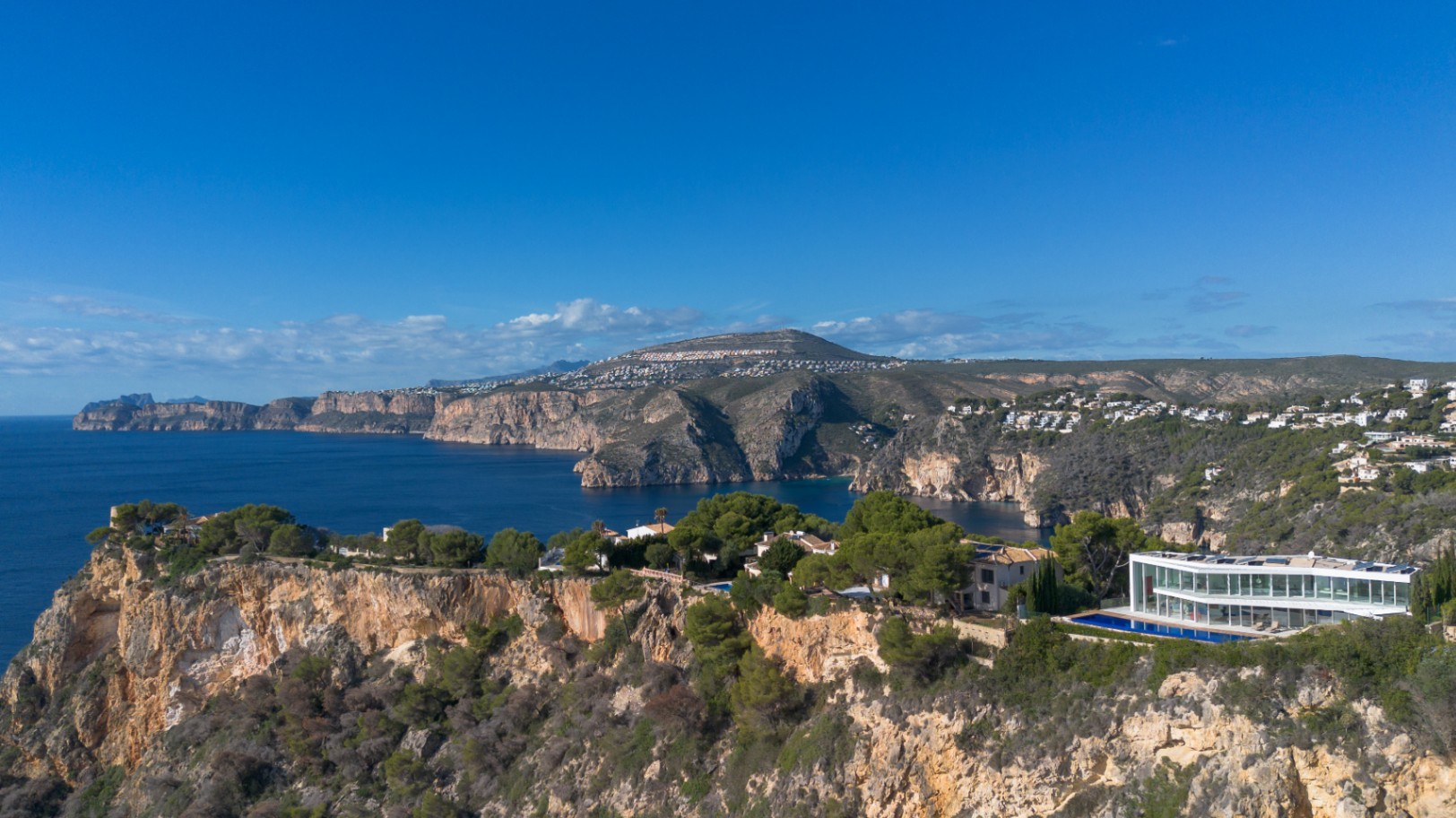 Aerial view of a contemporary glass-fronted villa perched on a cliffside, featuring a linear swimming pool and panoramic Mediterranean coastal views.