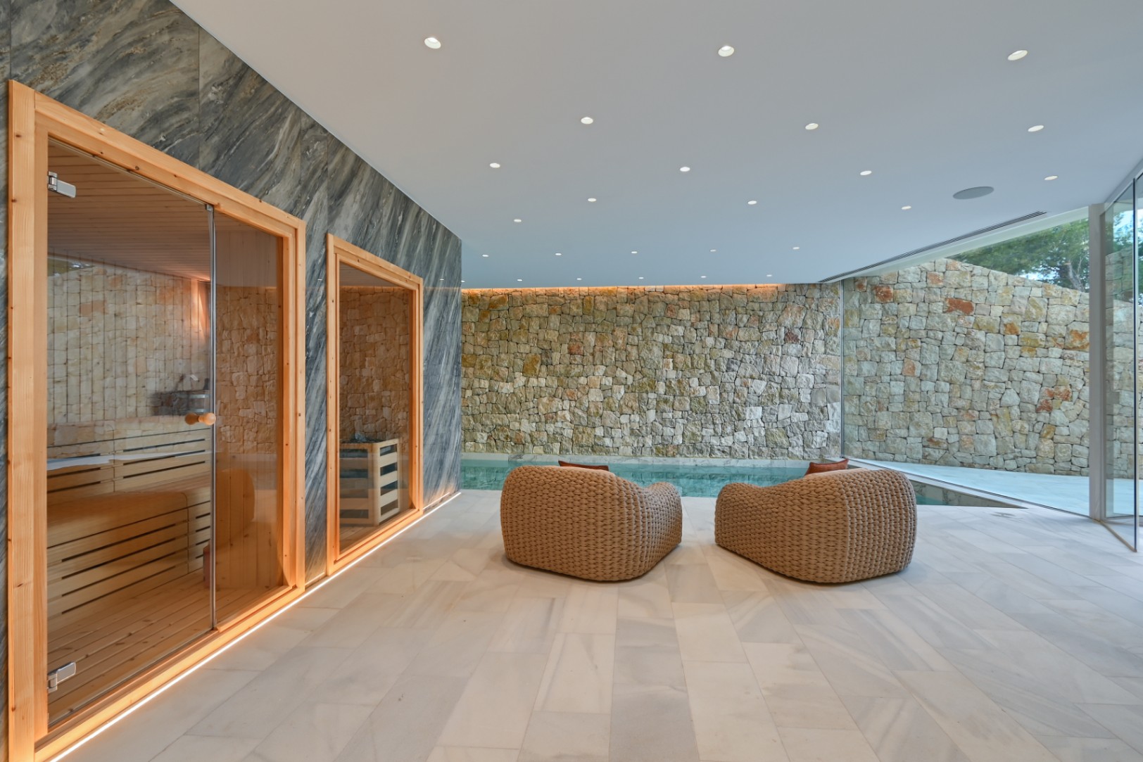 Indoor wellness suite featuring a wooden sauna with glass doors, woven lounge chairs, and a rustic stone wall adjacent to a private indoor pool.