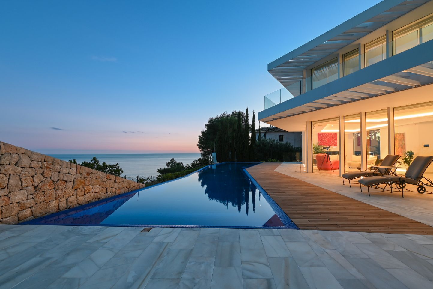 Infinity pool with wooden decking and stone paving, overlooking the sea at dusk. The villa features large glass facades and a natural stone wall.