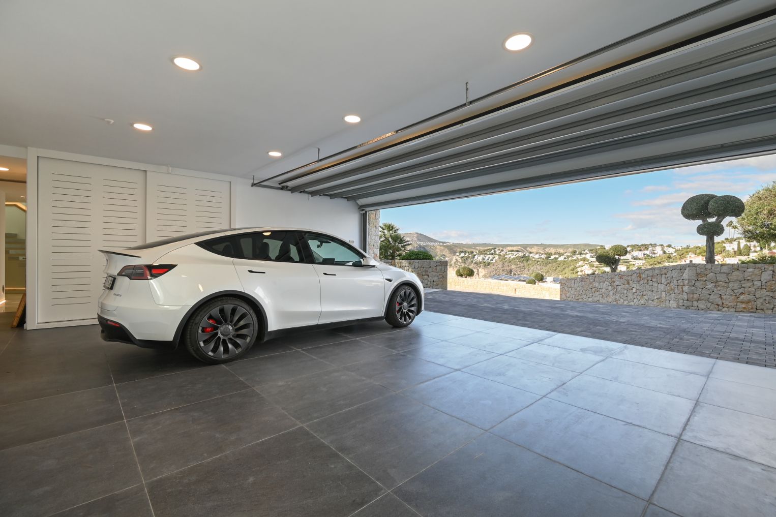 Modern garage featuring large grey floor tiles, recessed ceiling lights, and an open door overlooking a stone-paved driveway and valley views.