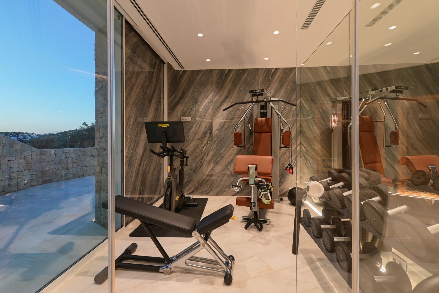 Modern home gym featuring Technogym equipment, marble-effect wall paneling, and floor-to-ceiling glass walls overlooking a stone terrace at dusk.