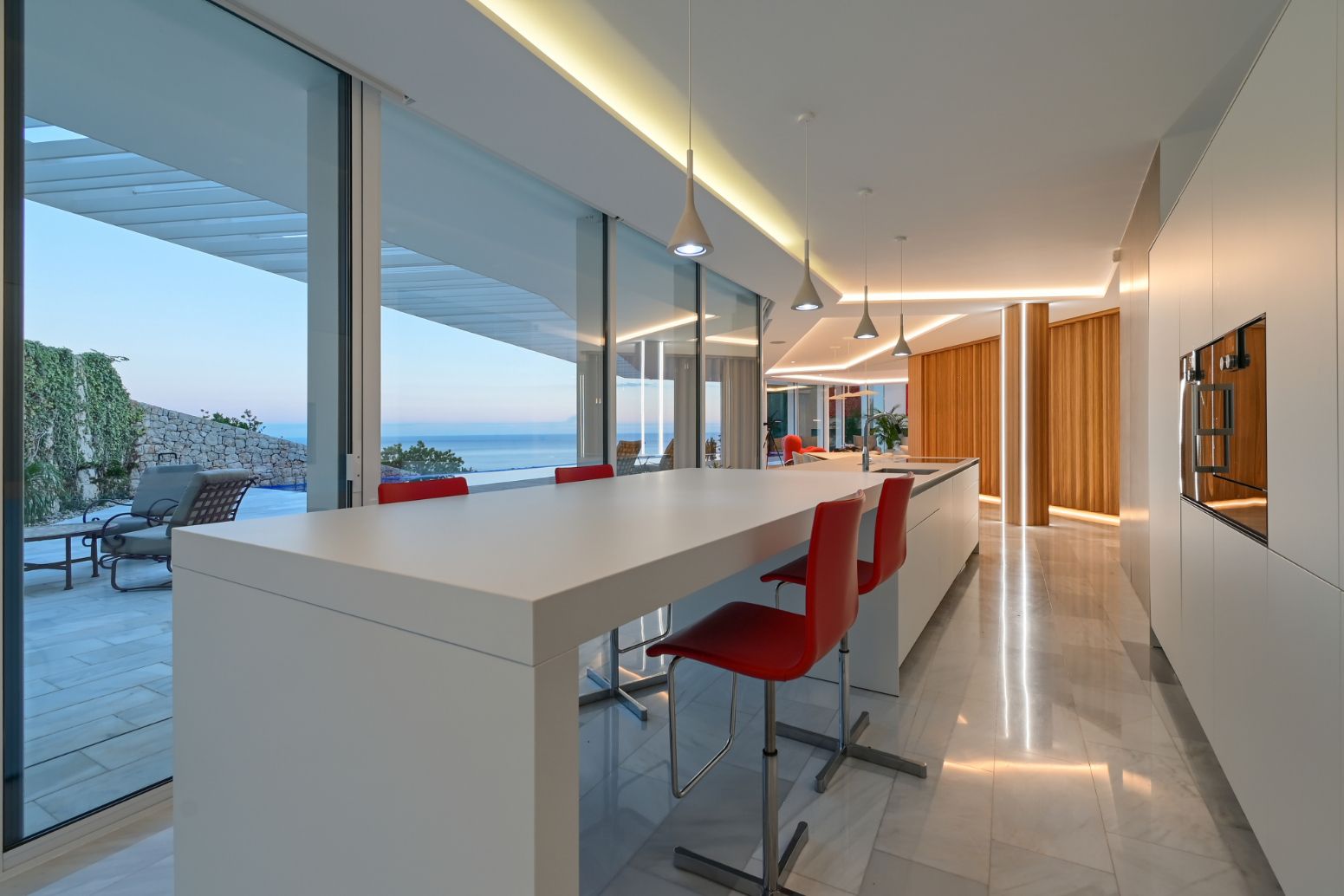 Modern kitchen with a white minimalist island and red stools, featuring floor-to-ceiling windows with sea views and integrated LED lighting.