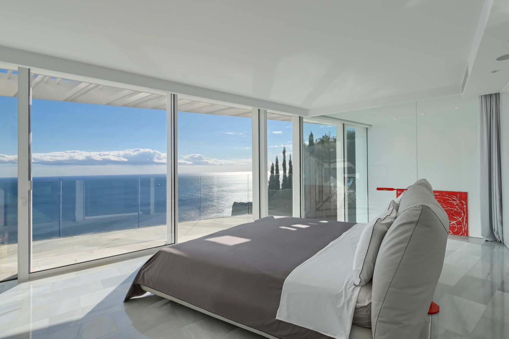 Modern master bedroom featuring floor-to-ceiling glass walls, white marble flooring, and panoramic Mediterranean Sea views from a private terrace.