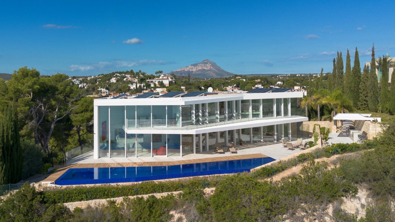 Modern two-story villa featuring floor-to-ceiling glass facades, a long infinity pool, solar panels, and panoramic views of the Montgó mountain.