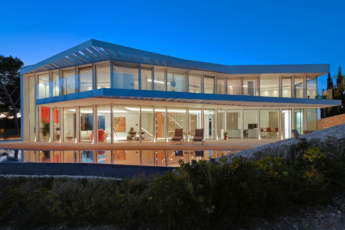 Modern two-story villa featuring floor-to-ceiling glass facades, an expansive terrace with a reflecting pool, and integrated lighting at dusk.