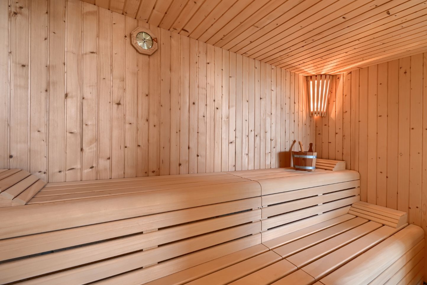 Sauna interior with light wood paneling, multi-level slatted benches, a corner light, and traditional wooden accessories like a bucket and ladle.