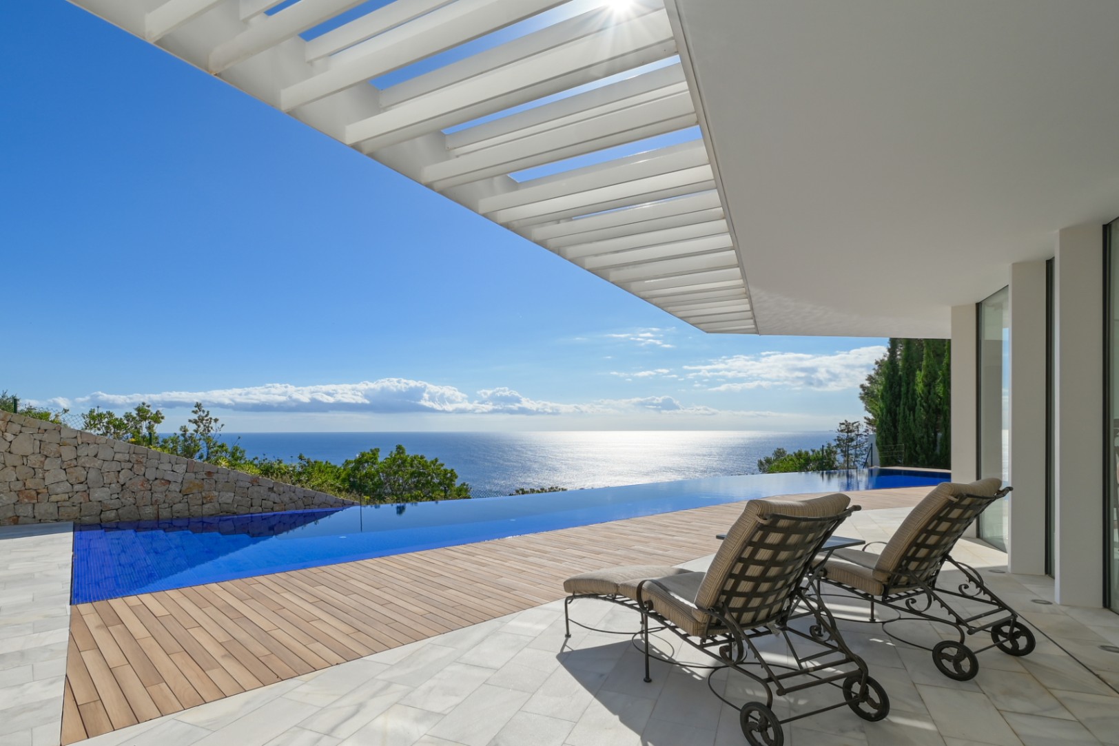 Terrace featuring an infinity pool with panoramic sea views, wooden decking, and marble flooring under a white architectural pergola with sun loungers.
