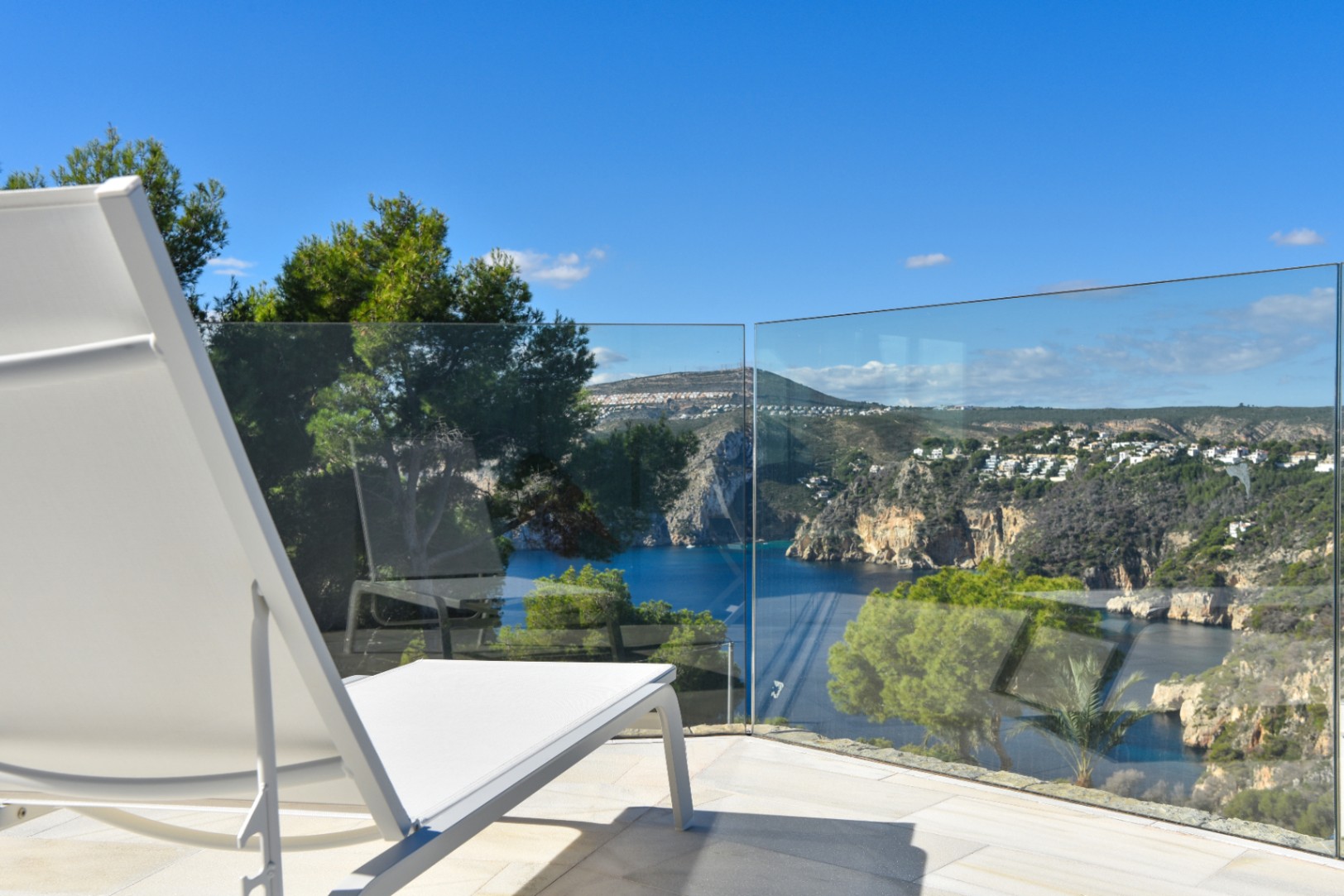 Terrace with white sun lounger and frameless glass railing, overlooking a Mediterranean bay and rugged coastal cliffs under a clear sky.